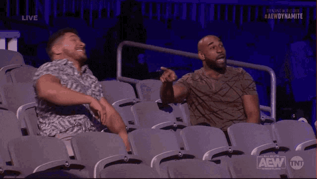 two men are sitting in a stadium laughing and watching a wrestling match