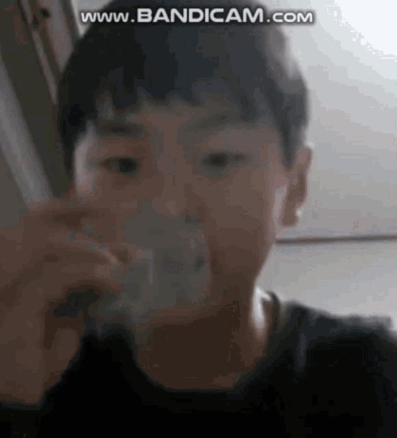 a young boy is drinking water from a glass while looking at the camera .