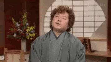 a man in a kimono is standing in front of a window in a room with flowers in a vase .