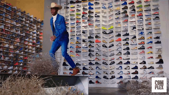 a man wearing a cowboy hat is standing in front of a wall of shoes with complex written on the bottom