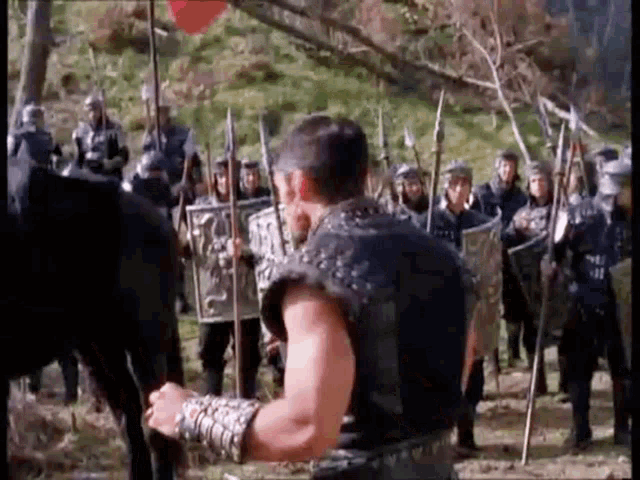 a man in a black vest is standing in front of a group of soldiers holding spears and shields .