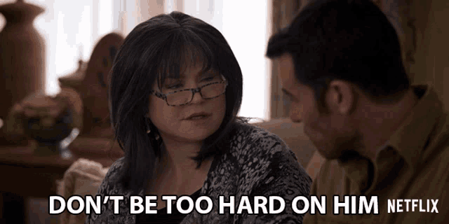 a man and a woman are sitting on a couch and the woman says " don 't be too hard on him "