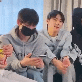 a man wearing a mask is looking at his phone while sitting next to another man .