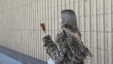 a woman in a leopard print coat is holding an ice cream bar in front of a brick wall
