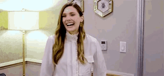 a woman in a white shirt is laughing in a room with a lamp and a picture on the wall .