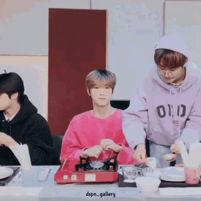 a group of young men are sitting at a table preparing food . one of the men is wearing a purple hoodie .