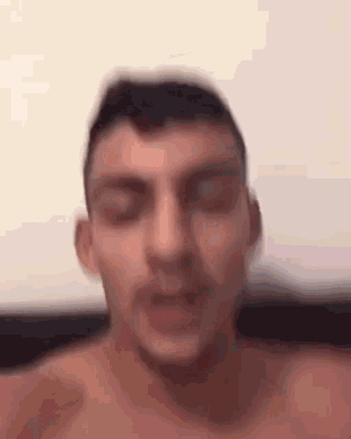 a man without a shirt is sitting on a bed with his eyes closed and his mouth open .