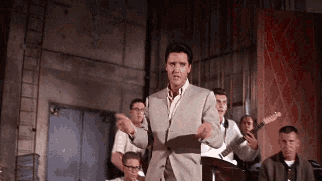 a man in a suit stands in front of a group of men playing guitars