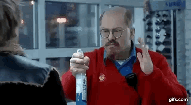 a man with glasses and a mustache is holding a spray bottle in his hand and talking to another man .