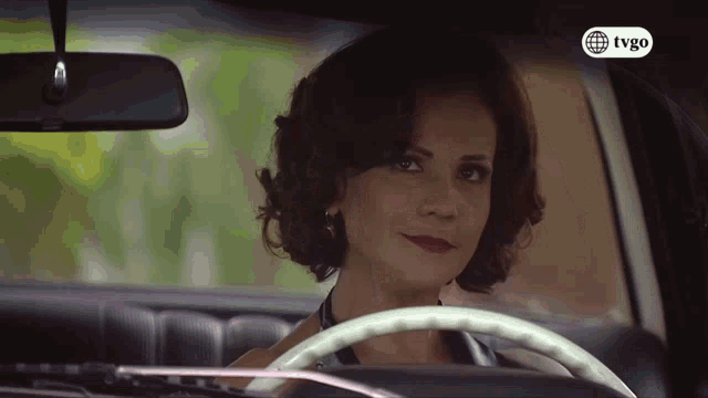 a woman is driving a car with a tvgo logo on the bottom right