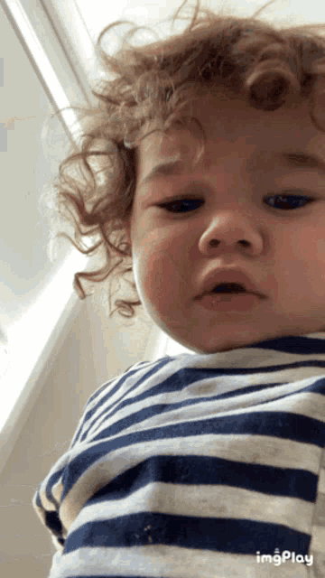 a baby with curly hair wearing a blue and white striped shirt with imgplay written below it