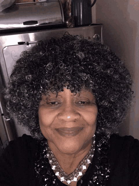 a woman wearing a gray afro wig and a necklace smiles for the camera