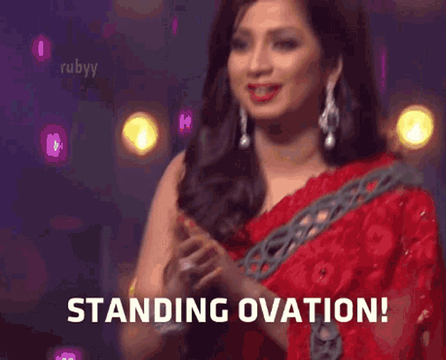 a woman in a red saree is standing in front of a crowd and says standing ovation !