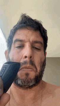 a shirtless man is shaving his beard with a trimmer .