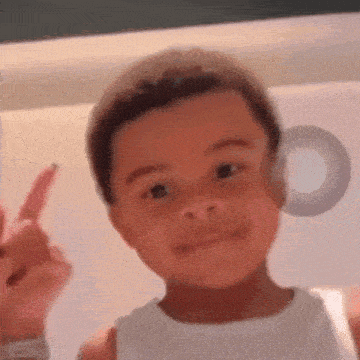a young boy in a white tank top is making a peace sign with his finger .