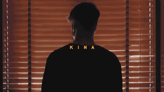 a man standing in front of a window with blinds and the word kina on his back