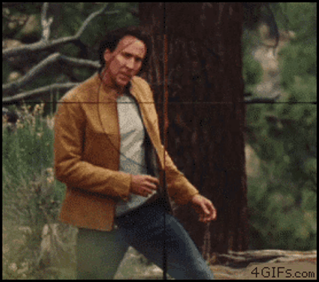 a man in a brown jacket is standing next to a tree with a sniper scope behind him