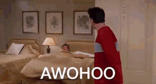 a man in a red shirt is standing in a bedroom next to a bed and a lamp .