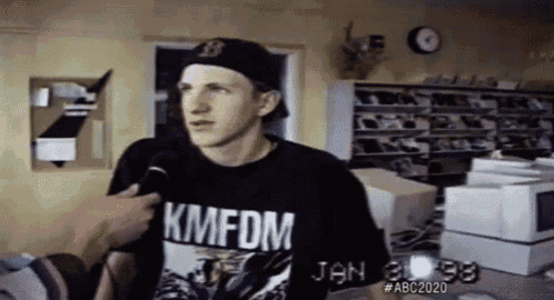 a man wearing a kmfdm t-shirt talks into a microphone