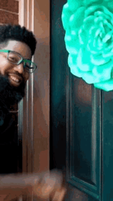 a man wearing glasses is standing in front of a door with a green flower hanging on it .