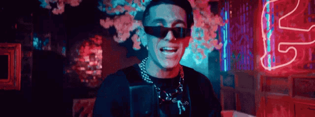 a man wearing sunglasses and a necklace is smiling in front of a neon sign that says n.