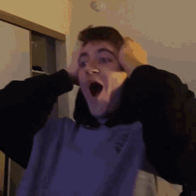 a young man in a hoodie is holding his hands to his head and making a surprised face .