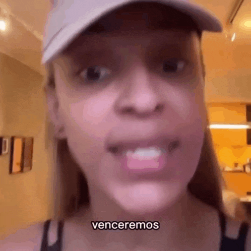 a woman wearing a hat and a black tank top is talking to the camera and says venceremos .