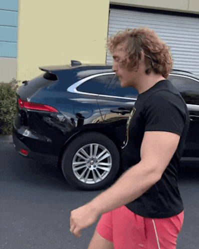 a man wearing red shorts and a black shirt is walking in front of a black car
