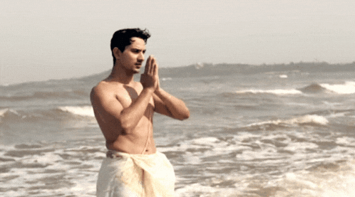 a shirtless man stands on the beach with his hands folded in prayer