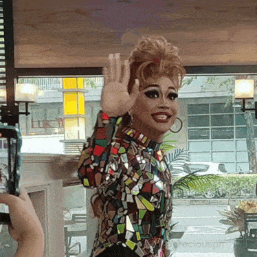 a drag queen waving her hand in front of a window with preciouspm written on the bottom right