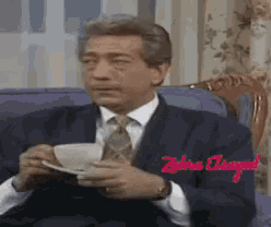 a man in a suit and tie sits in a chair drinking from a cup