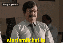 a man with glasses and a mustache is sitting in front of a sign that says starttamilchat.in .