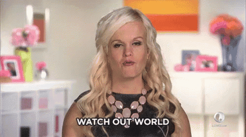a blonde woman is standing in a living room and says watch out world .