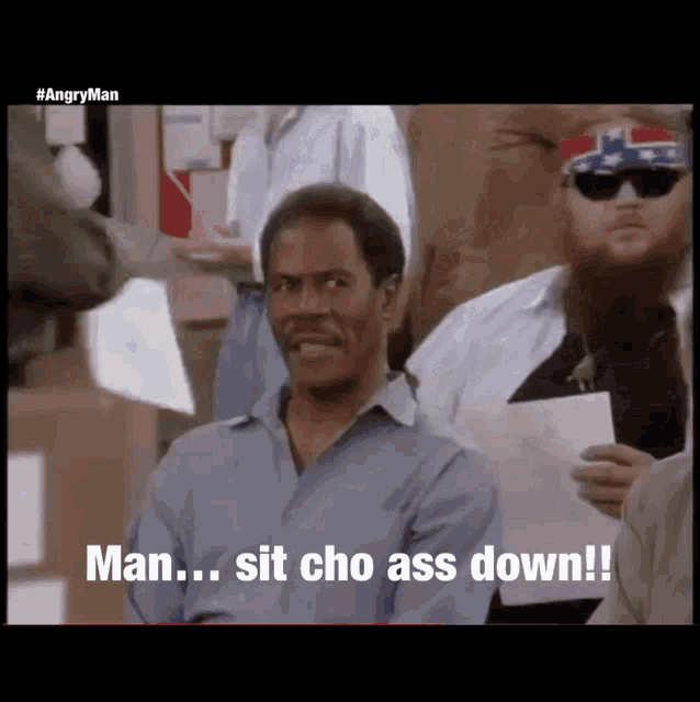 a man in a blue shirt is sitting in a crowd and says man sit cho ass down