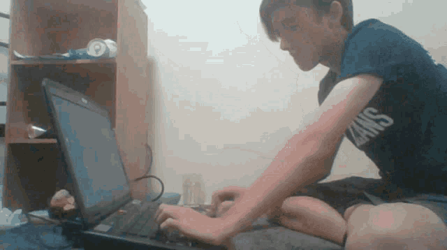 a boy wearing a blue vans shirt is typing on his laptop
