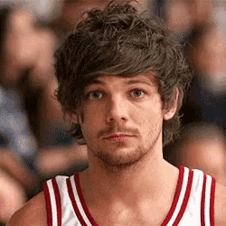 a young man with curly hair and a beard is wearing a red and white basketball jersey .
