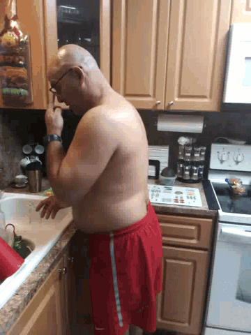 a shirtless man in red shorts is squatting on a counter in a kitchen