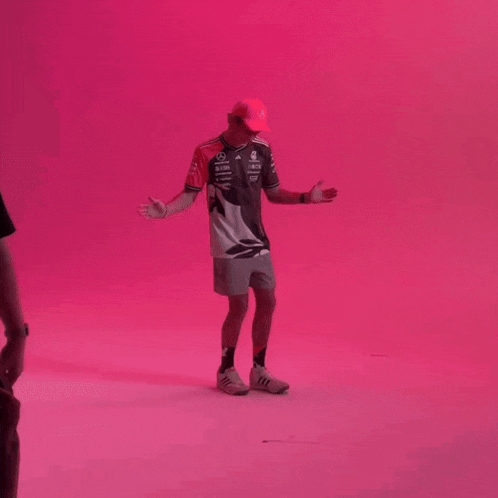 a man is standing in front of a pink wall with his arms outstretched and wearing a hat .