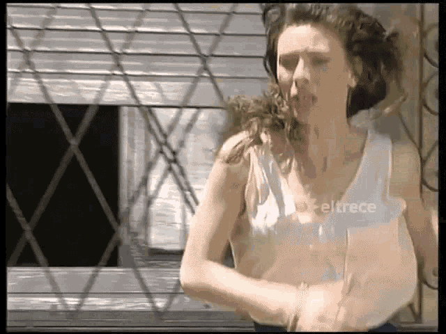 a woman in a white tank top is standing in front of a window with the word eltrece on the bottom right