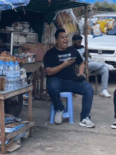 a man wearing a black shirt that says " regret is forgiven " sits on a blue stool