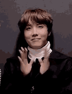 a young man wearing a black jacket and a white turtleneck is clapping his hands in front of a black wall .