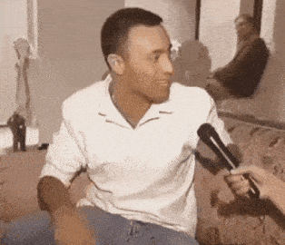a man in a white shirt is sitting on a couch being interviewed with a microphone .