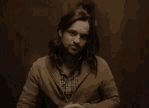 a man with long hair is sitting in front of a wooden wall with the word pas written on it .