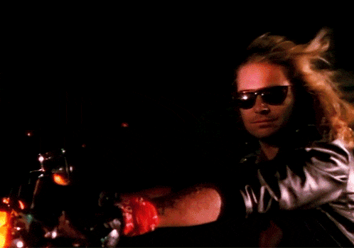 a man wearing sunglasses and a bandana on his wrist looks at the camera