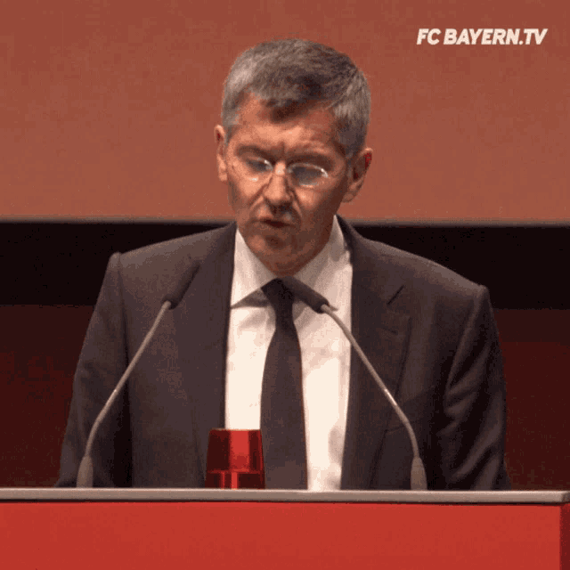 a man in a suit and tie is giving a speech at a podium with fc bayern.tv written above him