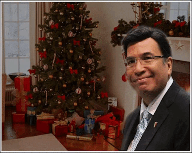 a man in a suit stands in front of a christmas tree and presents