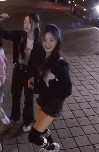 two girls are dancing on a sidewalk at night . one of the girls is wearing knee high socks .