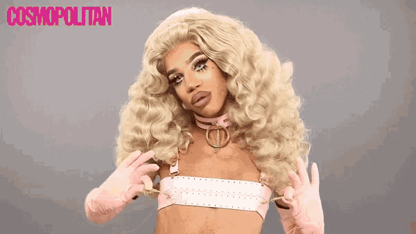 a woman in a blonde wig and pink gloves is posing for a picture in front of a cosmopolitan logo .