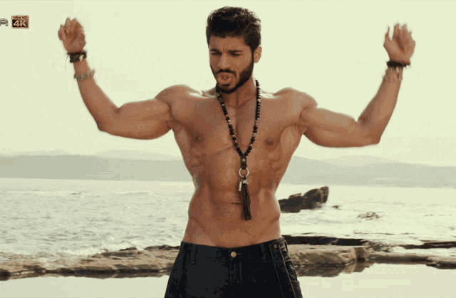 a man without a shirt is standing on a beach with his arms outstretched in front of a 4k screen