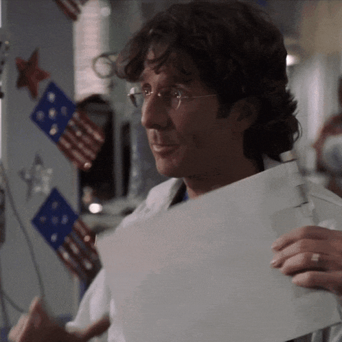 a man wearing glasses is holding a piece of paper in front of a wall with american flags on it